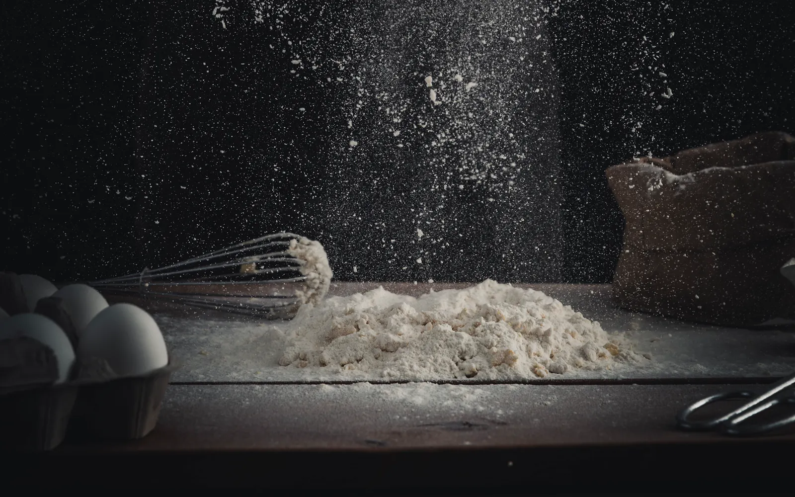 Flour being poured onto a wooden table beside eggs and a whisk